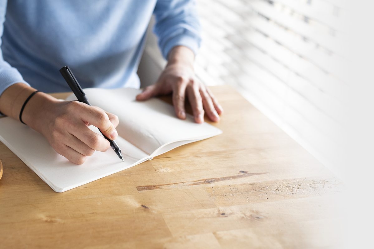 A person in a light blue sweater writing in an open notebook at a wooden desk