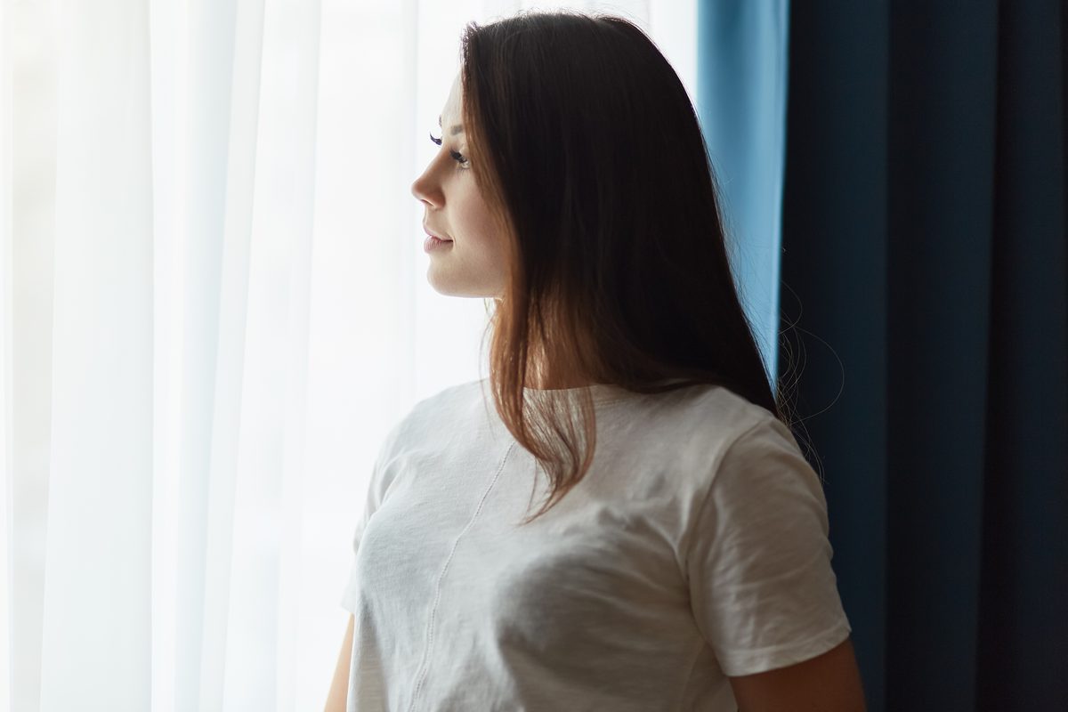 A woman in a white T-shirt standing by a window and looking out thoughtfully